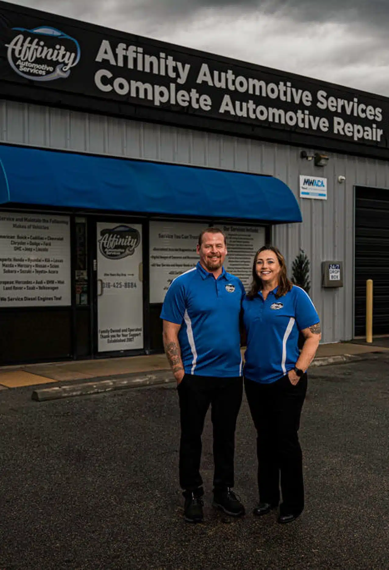 Two people wearing matching blue Affinity Automotive shirts stand and smile in front of the Affinity Automotive Services building under a cloudy sky.